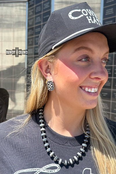 A woman wearing a black baseball cap with white embroidered text, a black and white beaded necklace, and a black and white checkered earring, photographed outdoors against a memorial wall available at Cowtown Bling N Things.