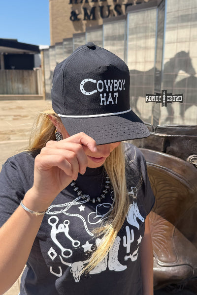 A woman wearing a black baseball cap with 'COWBOY HAT' embroidered in white on the front, photographed outdoors in front of a building with a cowboy boot and glass windows available at Cowtown Bling N Things.
