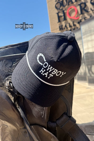 A black baseball cap with 'COWBOY HAT' embroidered in white on the front, displayed on a bronze cowboy statue outdoors. The background shows a sunny outdoor scene with a building and a blue sky available at Cowtown Bling N Things.