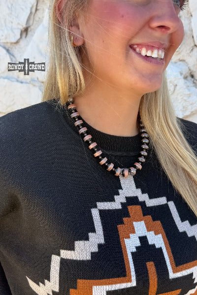 A close-up lifestyle shot of a woman wearing a beaded necklace with metallic rose gold and dark brown beads, paired with a black patterned knit sweater. The background is a light-colored stone wall available at Cowtown Bling N Things.
