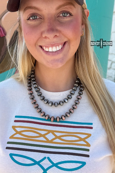 A close-up lifestyle shot of a smiling blonde woman wearing a white graphic t-shirt with colorful stripes and a double-strand beaded necklace. The necklace features metallic bronze and dark gray beads available at Cowtown Bling N Things.