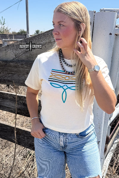 A woman wearing a white graphic t-shirt with a blue and orange geometric design, paired with light blue denim shorts, standing outdoors in front of a rustic wooden fence available at Cowtown Bling N Things.