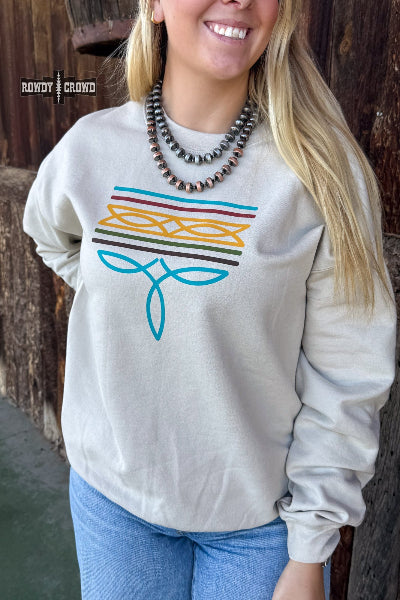 A woman wearing a light beige crewneck sweatshirt with a colorful graphic print on the front, paired with blue jeans and a beaded necklace, standing against a rustic wooden background available at Cowtown Bling N Things.