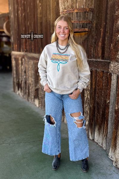 A woman stands in front of a rustic wooden wall wearing a cream sweatshirt with a colorful striped graphic and blue distressed jeans. The brand name 'Rowdy Crowd' is visible as an overlay on the left side available at Cowtown Bling N Things.