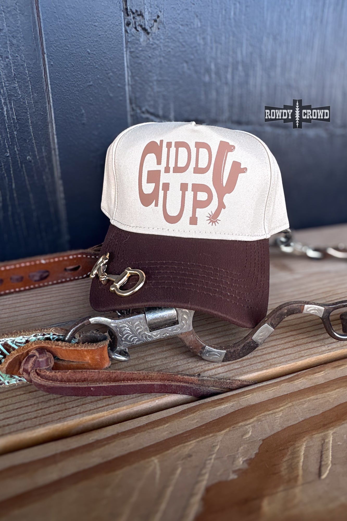 A two-tone trucker hat with a cream front panel and dark brown brim, featuring the words 'GIDDY UP!' in brown. The hat is styled on a wooden surface with leather and metal western accessories, against a dark blue paneled background available at Cowtown Bling N Things.