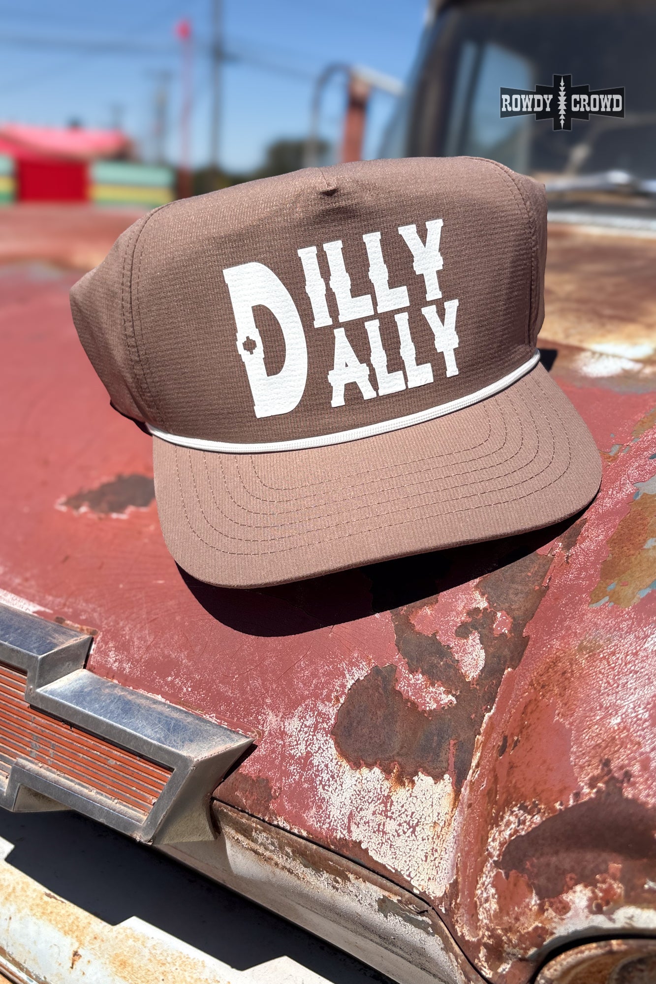 A brown trucker-style cap with 'DILLY DAILY' printed in white on the front, resting on the rusted hood of a vintage car outdoors. A 'Rusty Crowd' brand logo is visible in the top right corner.