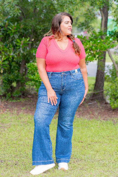 Woman wearing a pink top and blue jeans standing outdoors with greenery in the background