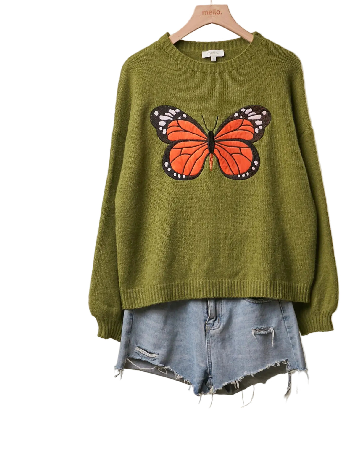 An olive green knit sweater with a large orange and black butterfly graphic on the front, styled on a wooden hanger with light-wash denim shorts against a neutral beige background.