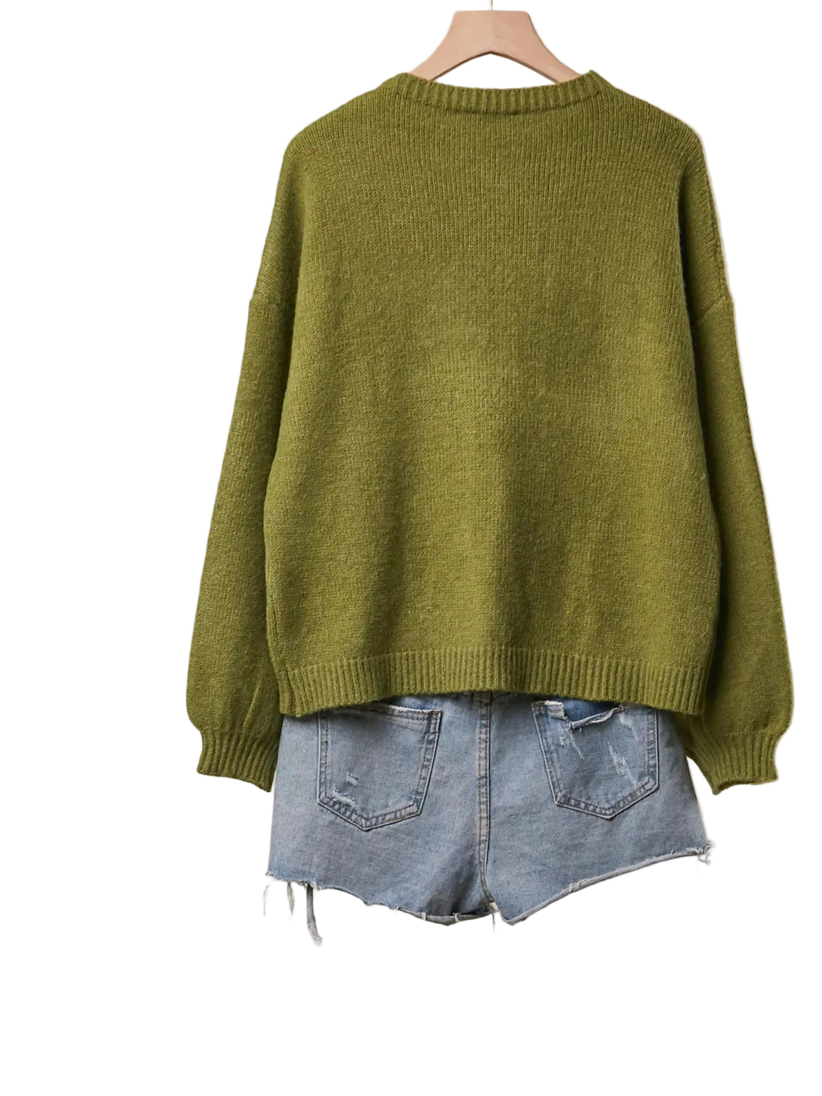 An olive green knit sweater hanging on a wooden hanger, styled with light-wash denim shorts against a beige wall, with a rustic wooden shelf and decorative items visible on the left.