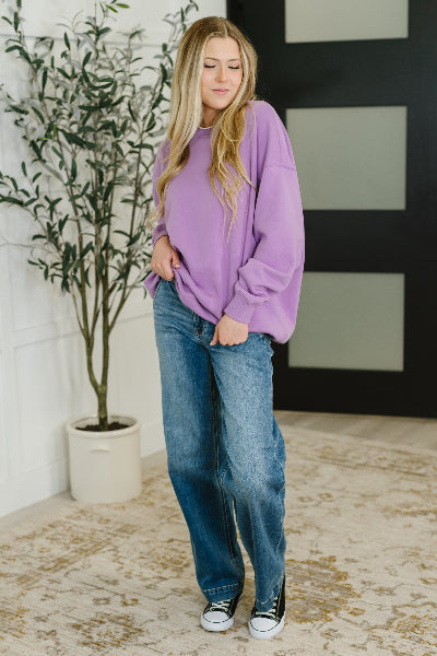 Woman wearing a purple sweater and blue jeans standing indoors with a plant and door in the background.