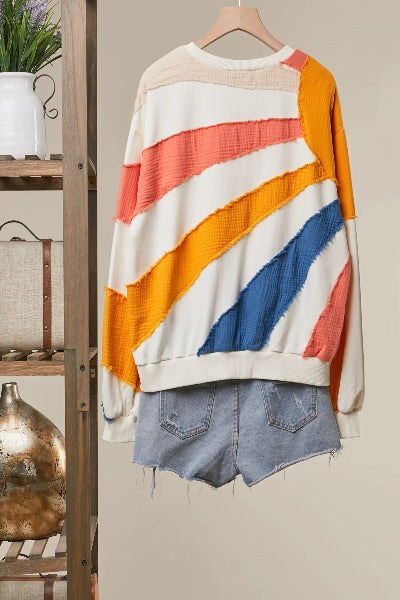 A long-sleeve crewneck sweater with diagonal textured stripes in cream, orange, coral, and blue, styled with light-wash denim shorts on a wooden hanger against a beige wall.