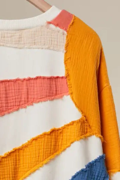 A close-up of a patchwork knit sweater featuring textured horizontal and diagonal stripes in mustard yellow, coral pink, cream, and blue on a white base.