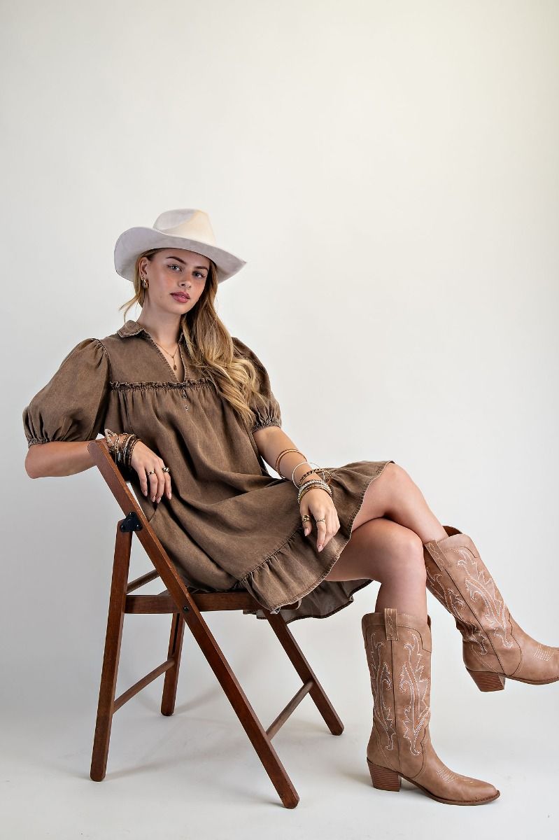 Woman in a brown dress and boots sitting on a wooden chair against a white background