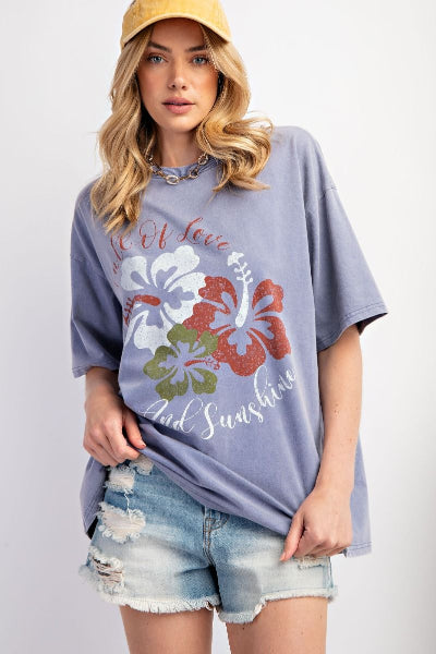 Woman wearing a light blue t-shirt with floral design and text, paired with denim shorts and a yellow cap.