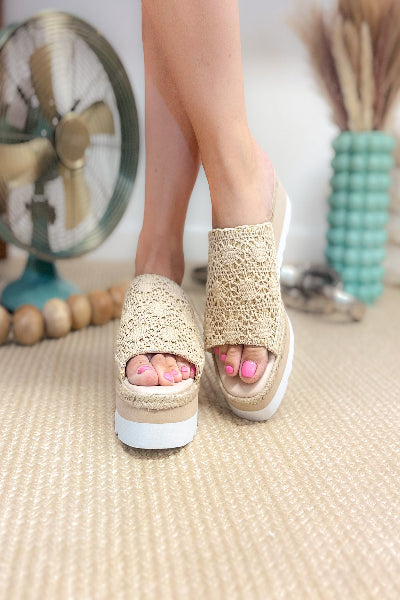 Beige sandals worn by a person on a textured floor with a fan and decorative items in the background.