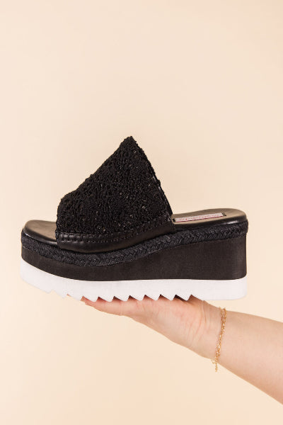 Black platform sandal held in a hand against a beige background