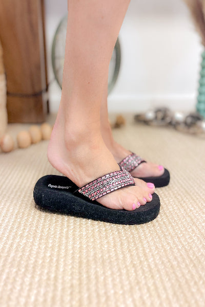 Black flip-flops with pink embellishments worn by a person on a beige carpet.